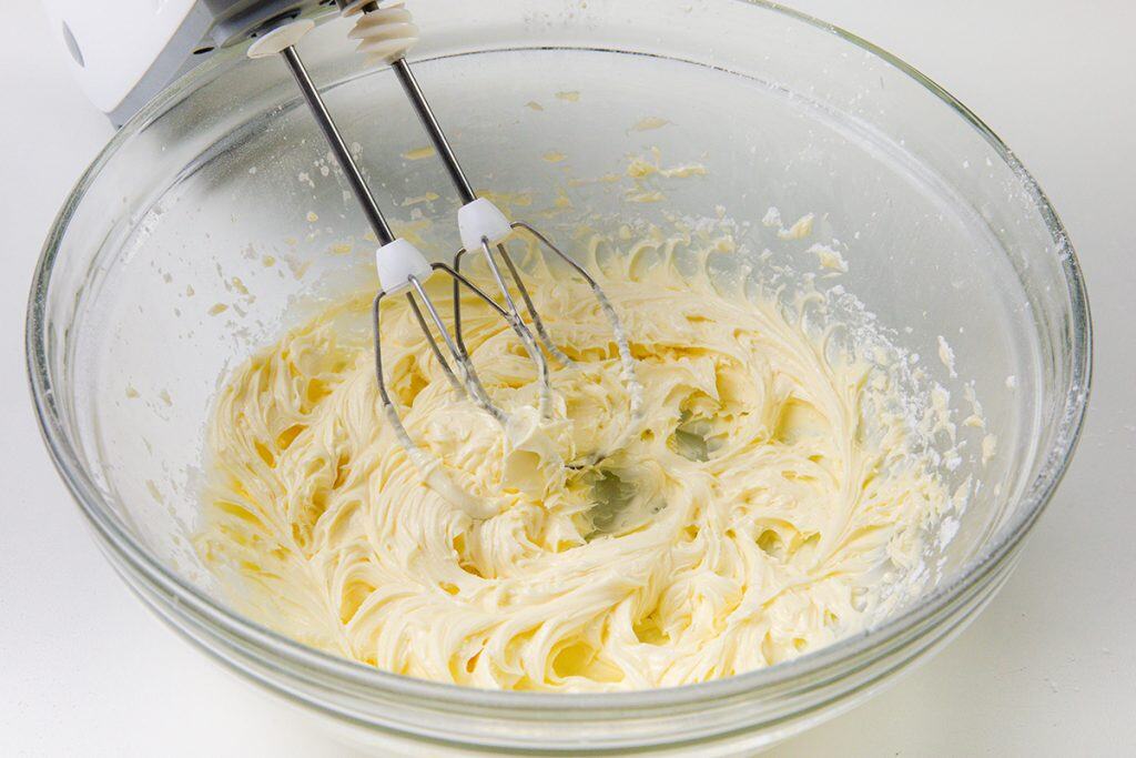 A glass bowl containing creamy, whipped butter and sugar—perfect for making snowball cookies with pecans—being mixed with an electric hand mixer on a white surface.