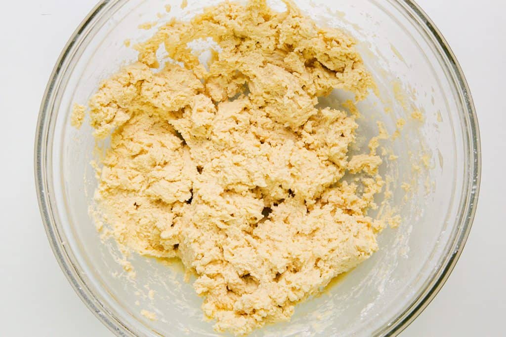 A glass bowl contains a rough, pale yellow dough mixture with a slightly crumbly texture, perfect for making classic Snowball Cookies or adding chopped pecans, viewed from above on a white surface.