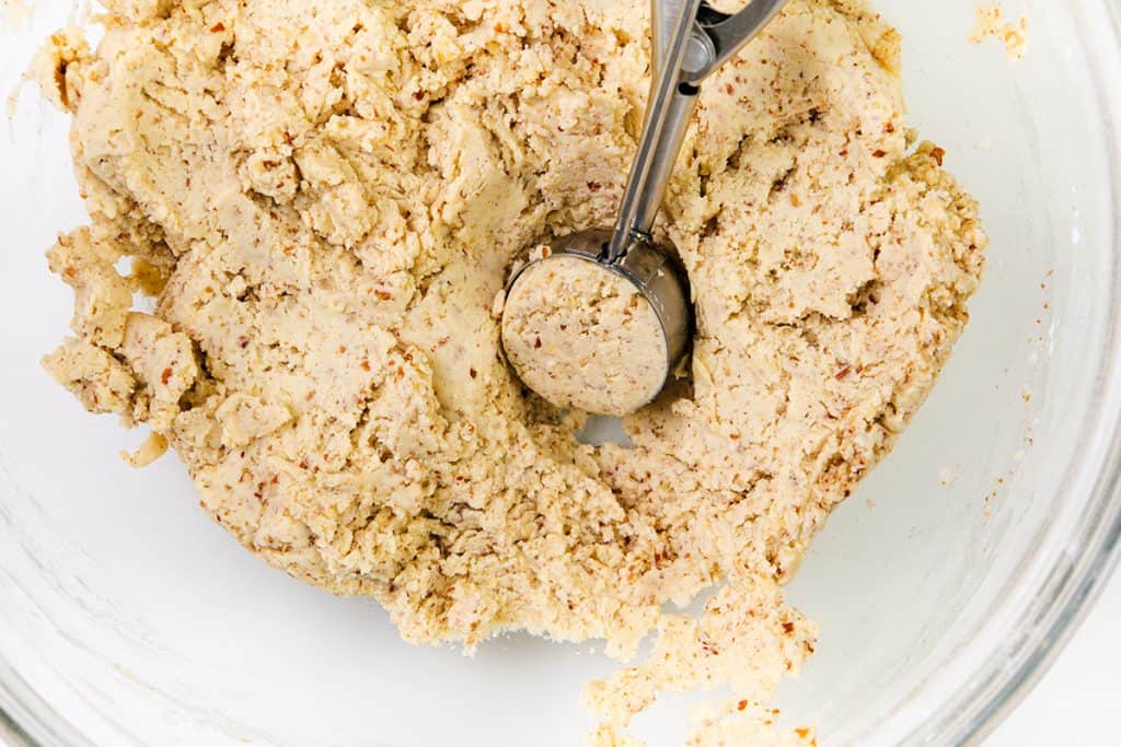 A metal cookie scoop pressing into a bowl of raw snowball cookie dough with visible pecans, inside a clear glass mixing bowl.