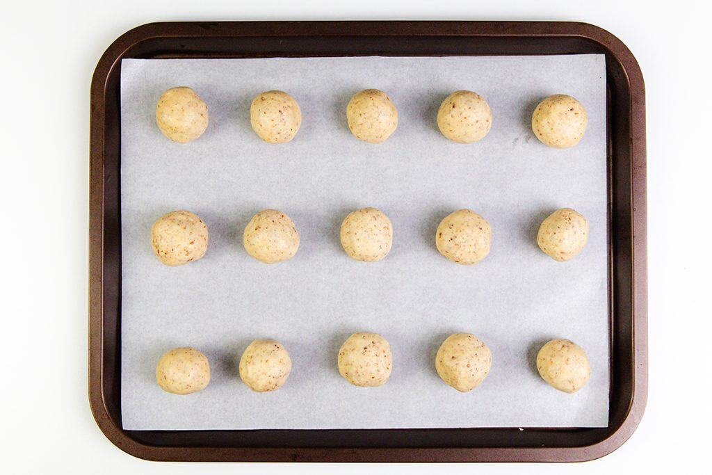 A baking tray lined with parchment paper holds fifteen evenly spaced, round Snowball Cookies arranged in a grid pattern, ready to be baked.