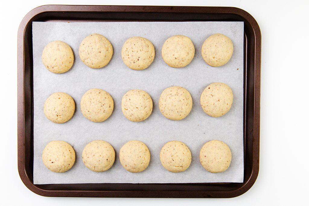 A baking tray lined with parchment paper holds sixteen evenly spaced Snowball Cookies arranged in a 4 by 4 grid pattern. The cookies are lightly golden, have a smooth surface, and are studded with chopped pecans.