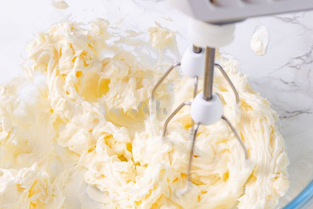 An electric hand mixer is whipping creamy, pale yellow champagne buttercream in a clear glass bowl on a marble countertop. The buttercream appears light and fluffy, with soft peaks forming&mdash;perfect for topping Champagne mini cupcakes.