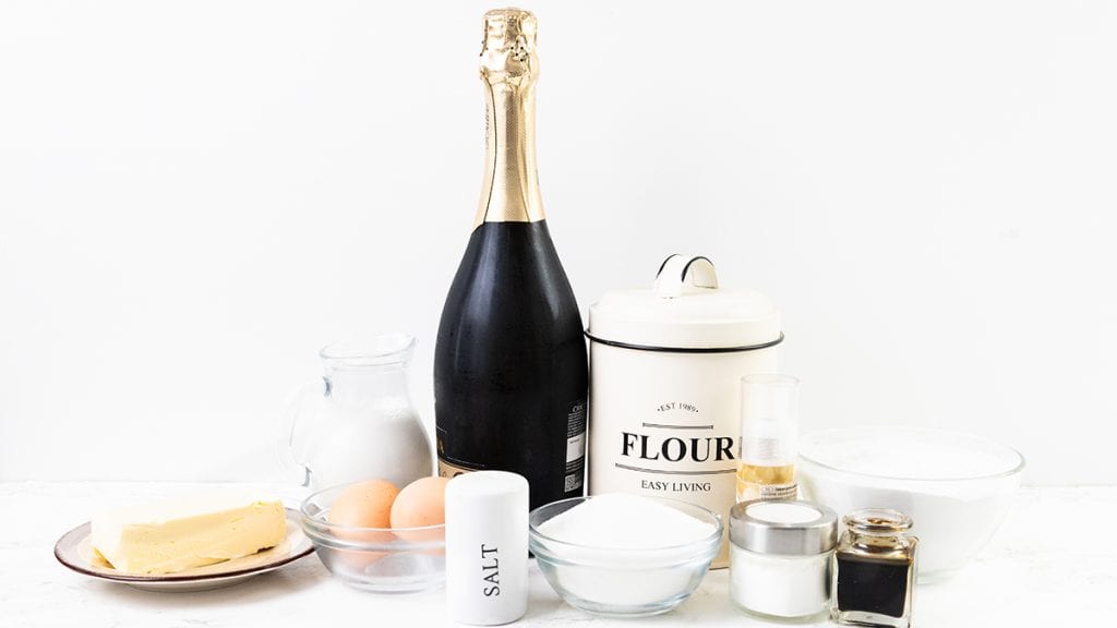 A selection of baking ingredients, perfect for making Champagne mini cupcakes, including a bottle of sparkling wine, butter, eggs, sugar, a flour canister, salt shaker, milk, and jars&mdash;all set on a white background.