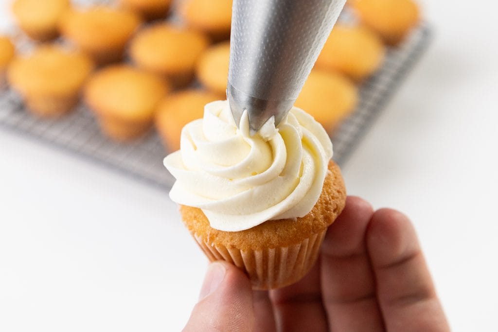 A hand holds a small cupcake while white champagne buttercream is piped onto it from a piping bag; more plain mini cupcakes are visible on a cooling rack in the background.