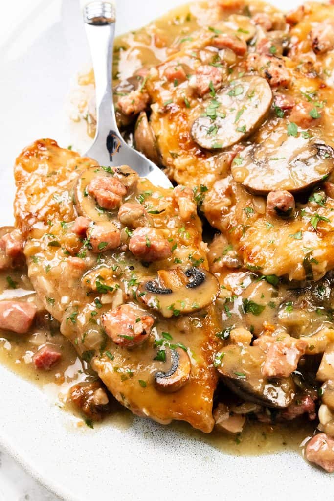 A close-up of Chicken Marsala on a plate, featuring golden-brown chicken breasts topped with a creamy marsala sauce, sliced mushrooms, diced pancetta, and sprinkled chopped parsley. A fork rests beside the chicken.