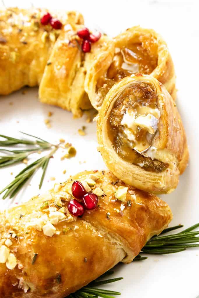 A close-up of golden, flaky puff pastry slices filled with creamy brie and rich fig jam, topped with chopped nuts and pomegranate seeds. Garnished with rosemary sprigs, this Puff Pastry Christmas Wreath sits beautifully on a white plate.