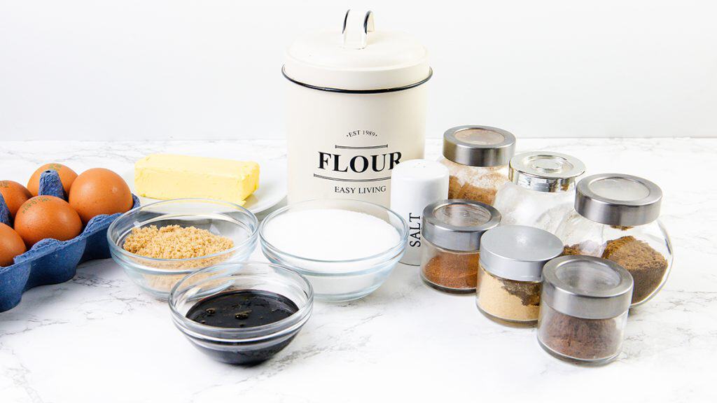 Berbagai bahan kue untuk Gingersnap Cookies di atas meja putih, termasuk telur, mentega, tepung, gula, garam, molase, dan berbagai macam bumbu dan bubuk dalam toples dan mangkuk.