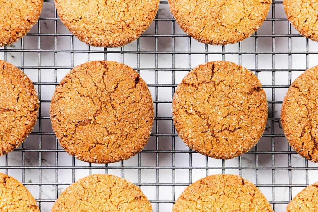 Round, golden-brown Gingersnap Cookies with crackled tops are arranged neatly on a metal cooling rack. The surface beneath the rack is white with light gray marbling.