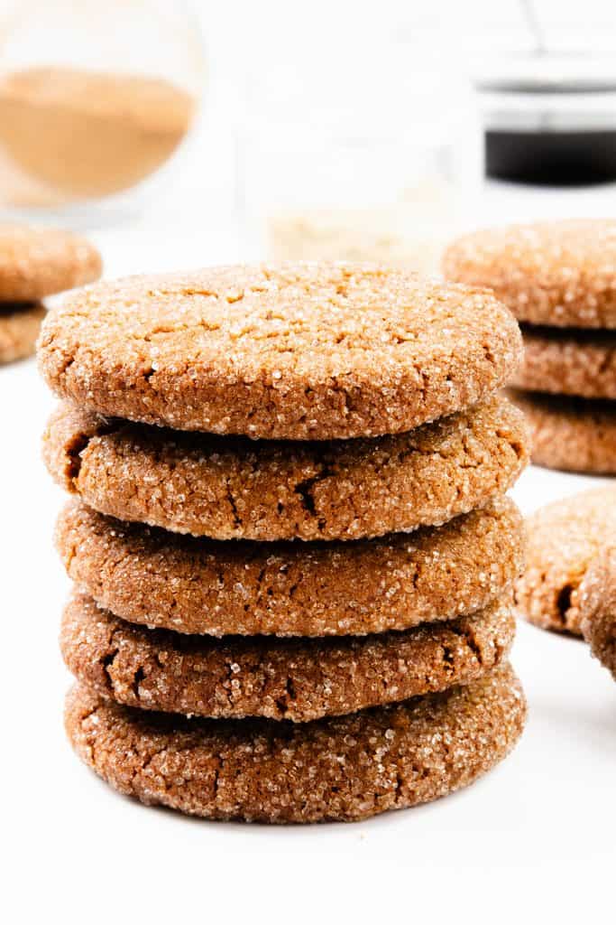 Tumpukan enam kue jahe berlapis gula ditampilkan di permukaan putih, dengan lebih banyak kue dan stoples berisi bahan-bahan yang diburamkan di latar belakang.