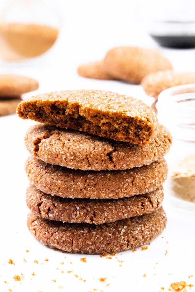 Setumpuk empat kue jahe berlapis gula dengan kue kelima dipecah menjadi dua dan diletakkan di atasnya. Remah-remah dan toples bahan-bahan terlihat di latar belakang permukaan putih.