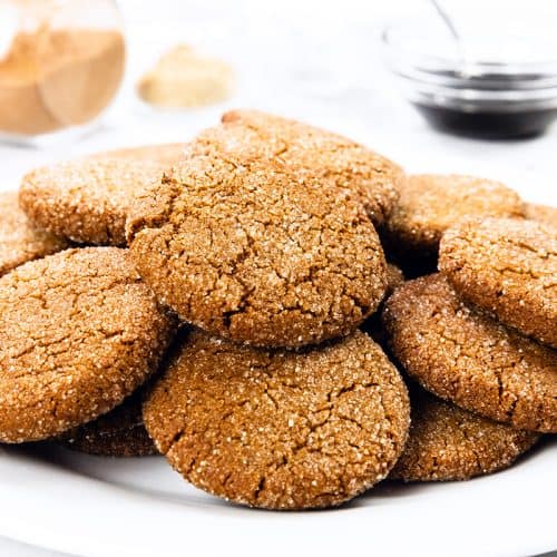 Piring putih berisi Kue Gingersnap berwarna coklat keemasan dan dilapisi gula. Pada latar belakang buram, toples kaca berisi kayu manis, semangkuk kecil sirup berwarna gelap, dan beberapa remah kue terlihat di permukaan putih.
