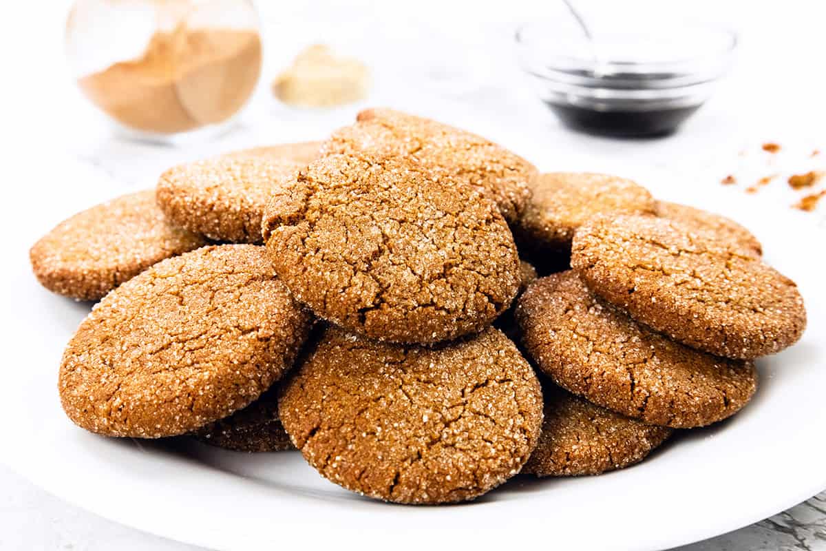 Piring putih berisi Kue Gingersnap berwarna coklat keemasan dan dilapisi gula. Pada latar belakang buram, toples kaca berisi kayu manis, semangkuk kecil sirup berwarna gelap, dan beberapa remah kue terlihat di permukaan putih.