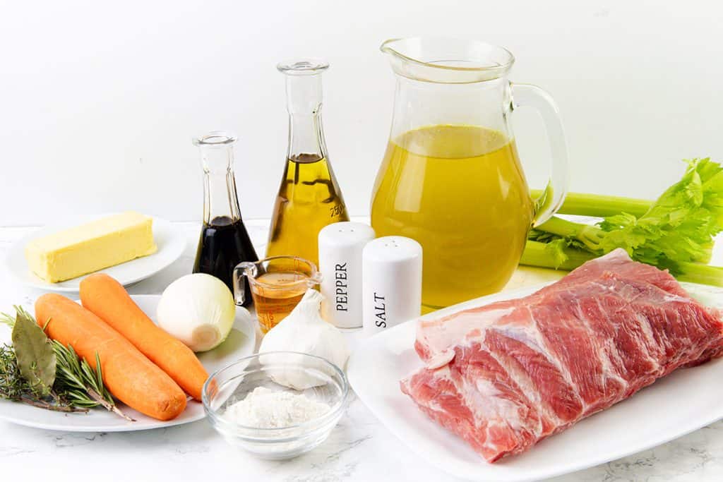 A variety of ingredients for a delicious Pork Gravy Recipe are arranged on a white surface, including raw pork ribs, carrots, onion, celery, garlic, butter, flour, herbs, bay leaves, broth in a pitcher, balsamic vinegar, oil, and seasonings.