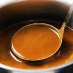 A metal ladle scoops rich pork gravy from a stainless steel pot filled with smooth, glossy gravy—perfect for any make-ahead gravy needs.