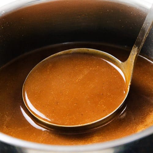 A metal ladle scoops rich pork gravy from a stainless steel pot filled with smooth, glossy gravy—perfect for any make-ahead gravy needs.