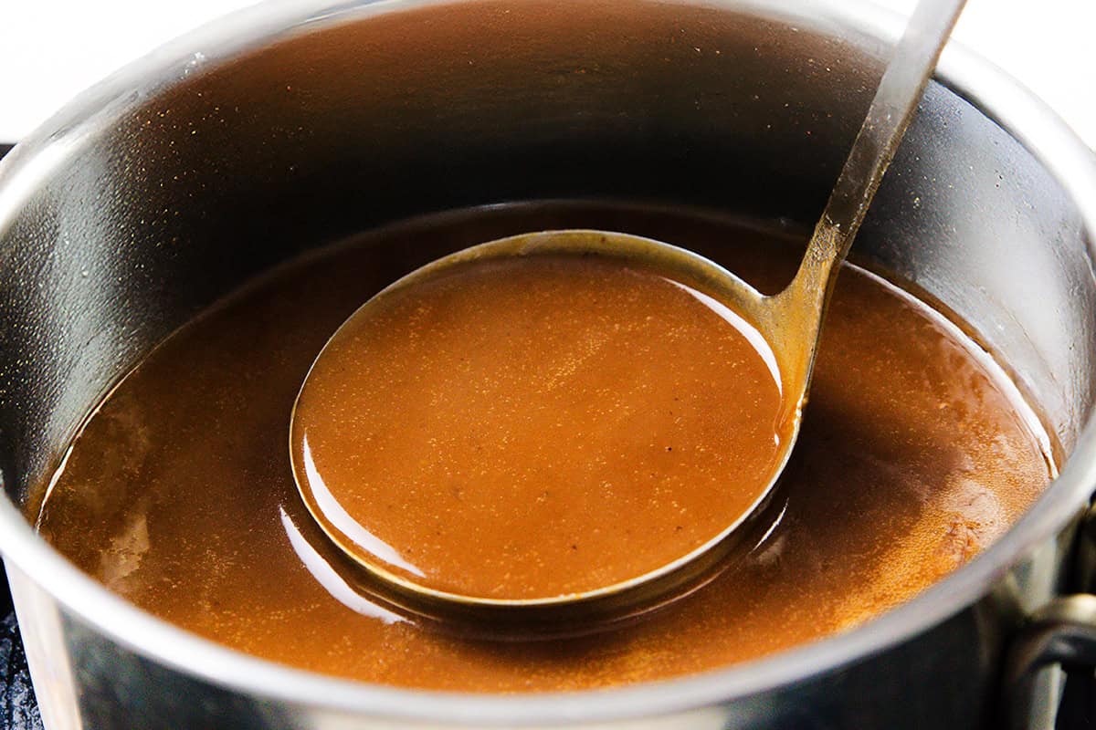 A metal ladle scoops rich pork gravy from a stainless steel pot filled with smooth, glossy gravy—perfect for any make-ahead gravy needs.