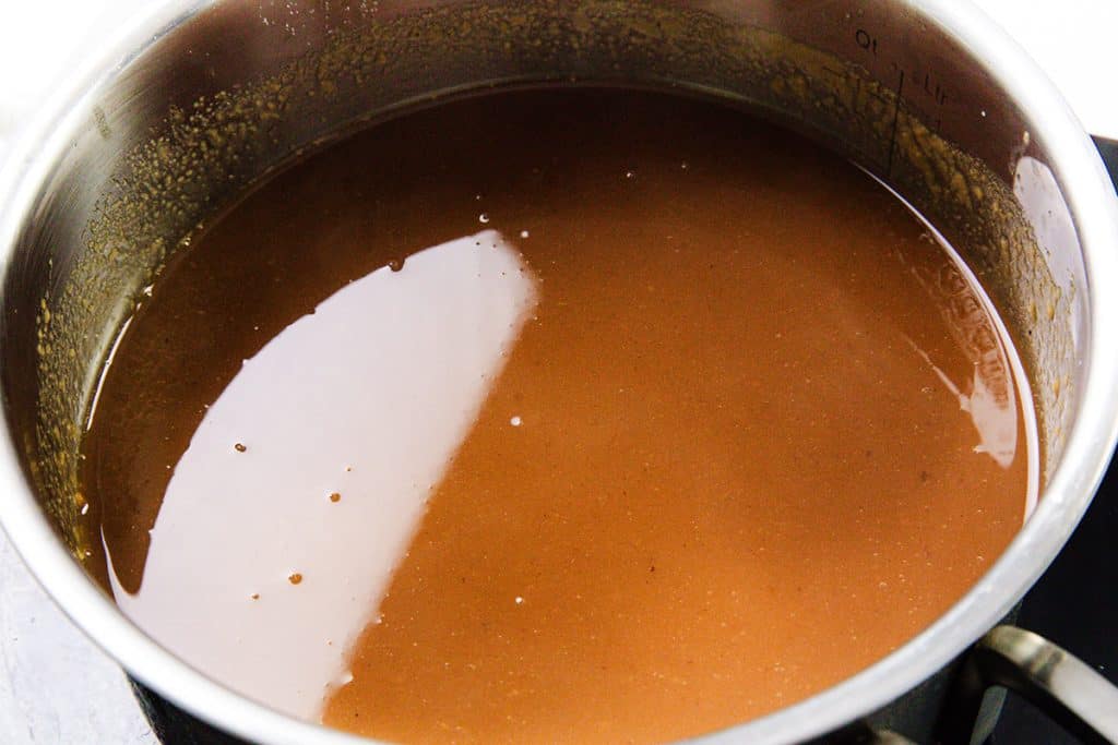 A stainless steel pot filled with smooth, brown pork gravy is heating on a stovetop, with some bubbles and a slight reflection visible on the surface—perfect for your favorite make-ahead pork gravy recipe.