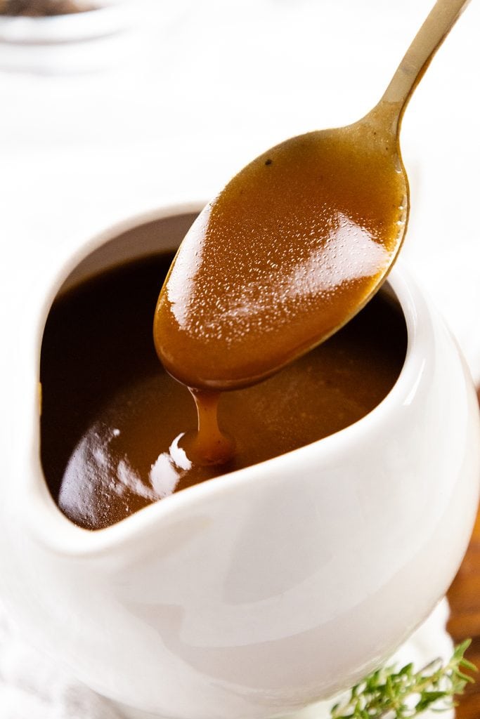 A close-up of golden-brown pork gravy being poured from a spoon into a white ceramic gravy boat, with its smooth and glossy texture visible—perfect for any pork gravy recipe or make-ahead meal.