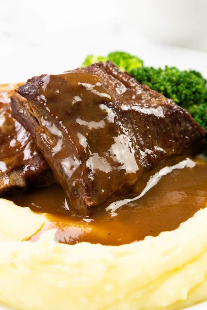A piece of braised beef covered in rich make-ahead gravy sits on a bed of creamy mashed potatoes, with steamed broccoli in the background.
