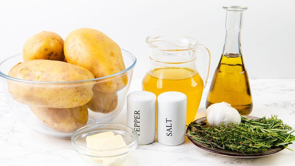 A bowl of raw potatoes ready for crispy potatoes, a glass jug of broth, a glass bottle of oil, fresh garlic and herbs, a dish of butter, and salt and pepper shakers on a white countertop.