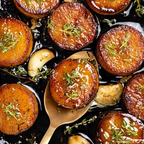 Golden, caramelized rounds of melting potatoes topped with fresh herbs sit in a pan with roasted garlic cloves and sprigs of thyme; a gold spoon lifts one crispy potato slice.
