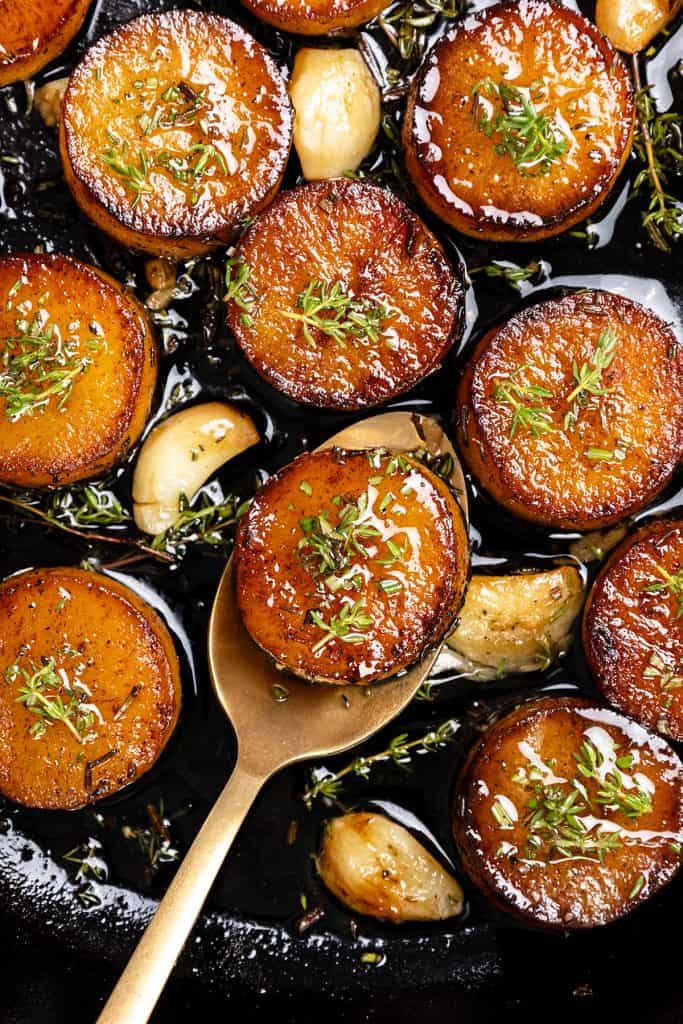 Golden, caramelized rounds of melting potatoes topped with fresh herbs sit in a pan with roasted garlic cloves and sprigs of thyme; a gold spoon lifts one crispy potato slice.