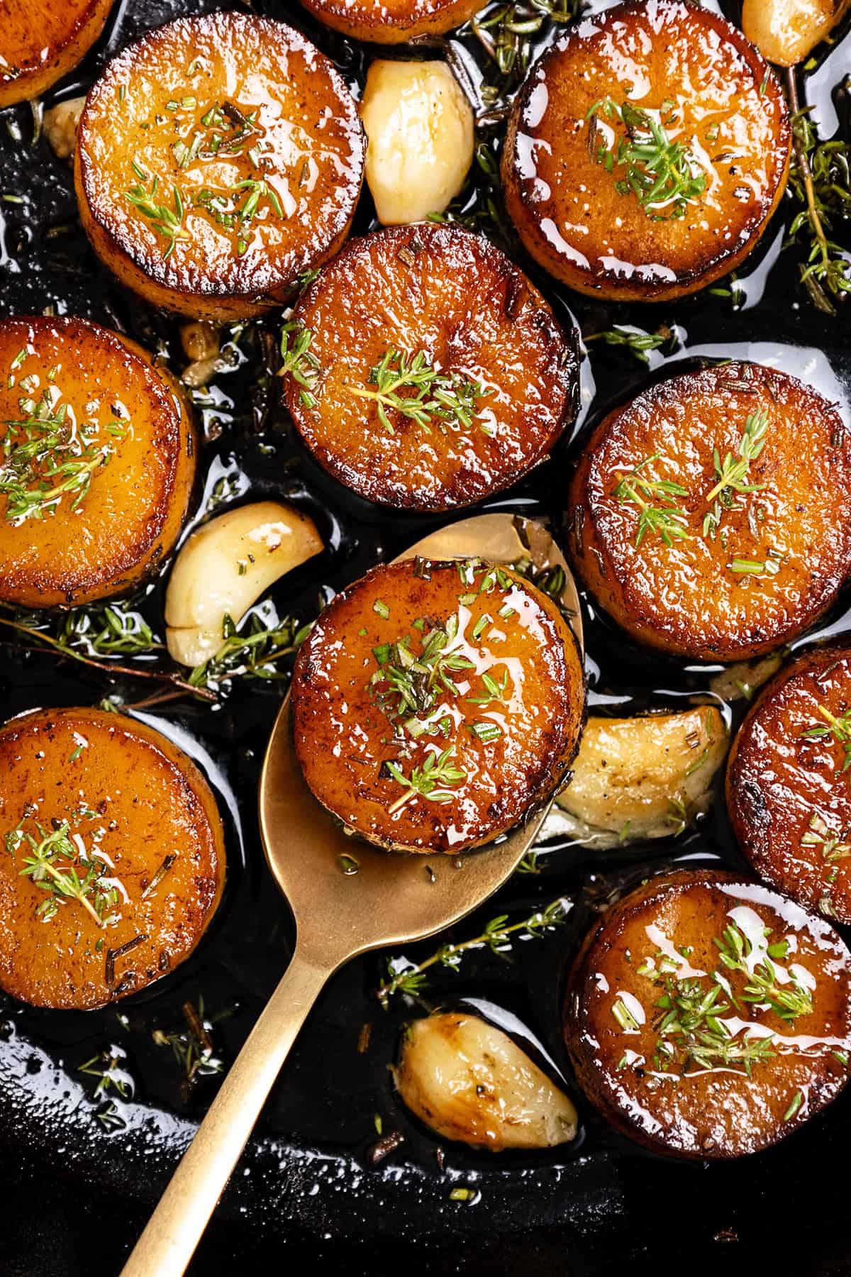 Golden, caramelized rounds of melting potatoes topped with fresh herbs sit in a pan with roasted garlic cloves and sprigs of thyme; a gold spoon lifts one crispy potato slice.