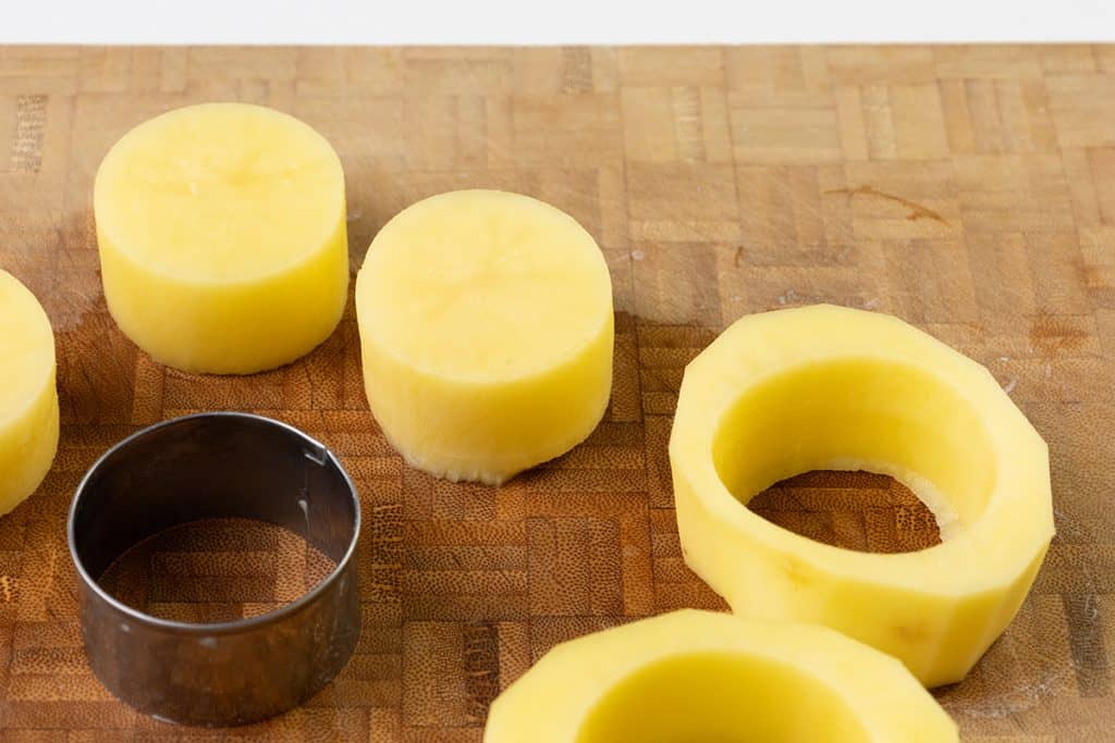 Thick slices of peeled potato are shown on a wooden cutting board, some with circular centers removed—perfect for a crispy potatoes recipe. A metal round cutter rests nearby, ready to help craft your next Melting Potatoes dish.