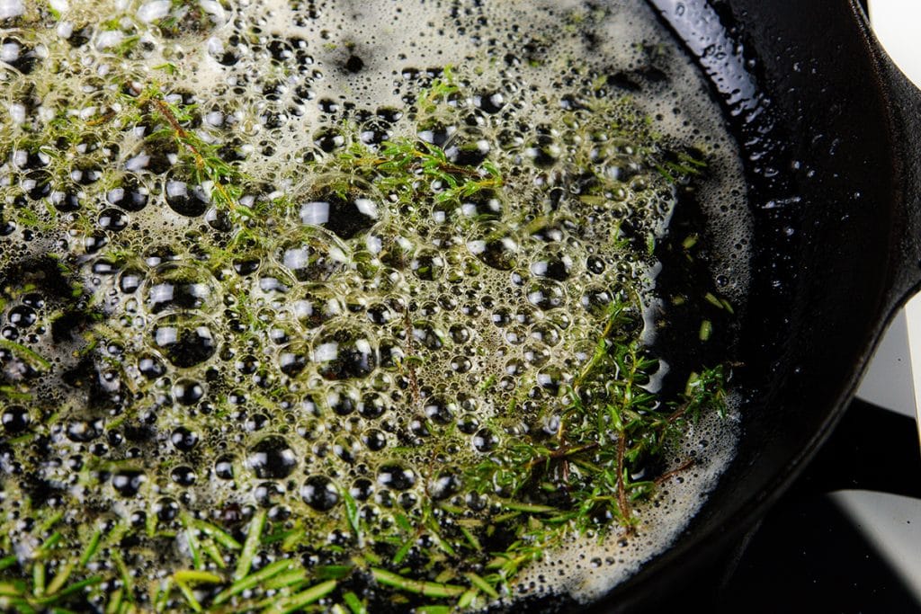 Close-up of a cast iron skillet with bubbling melted butter and fresh green herbs, like thyme and rosemary, perfect for creating Melting Potatoes—a delicious potato recipe featuring golden, crispy potatoes sautéed to perfection.