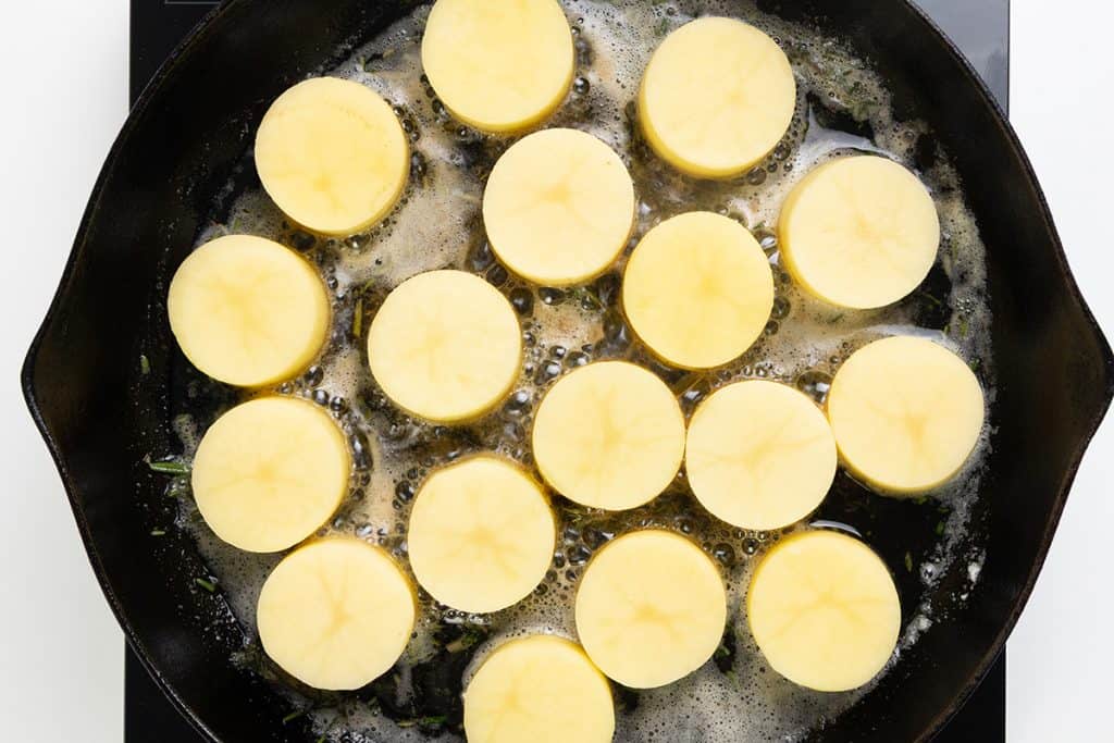 Sliced rounds of yellow potatoes, prepared in the style of Melting Potatoes, are cooking in a black skillet with bubbling butter. This delicious side dish is arranged in a single, even layer and seen from above.