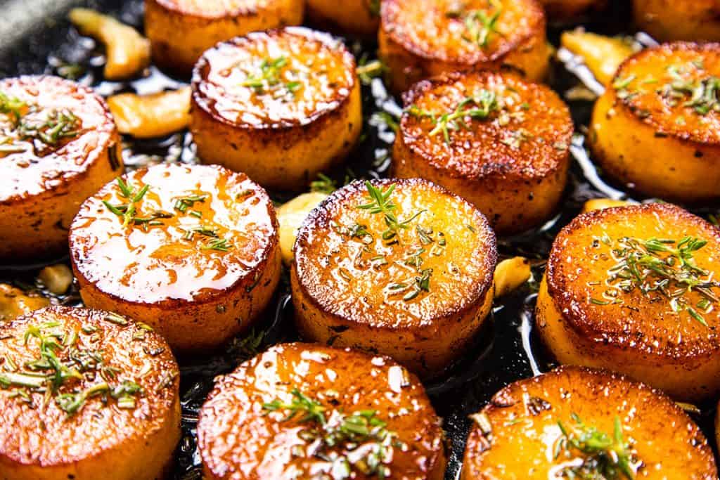 Golden brown Melting Potatoes with crisp edges sizzle in a pan of glistening butter and visible garlic, garnished with fresh herbs. This irresistible potato recipe delivers glossy, caramelized surfaces and ultimate flavor.