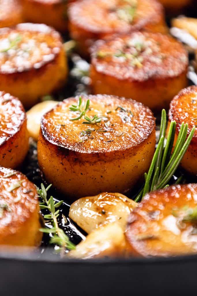 Golden brown, crispy roasted potatoes—perfect for any potato recipes—are garnished with fresh rosemary and thyme, surrounded by roasted garlic cloves in a skillet.