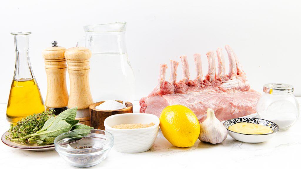 A raw rack of lamb sits on a white surface surrounded by olive oil, fresh herbs, a lemon, garlic, mustard, breadcrumbs, salt, pepper grinders, a bowl of water, and a small bowl of sugar—perfect ingredients for a savory garlic herb crust.