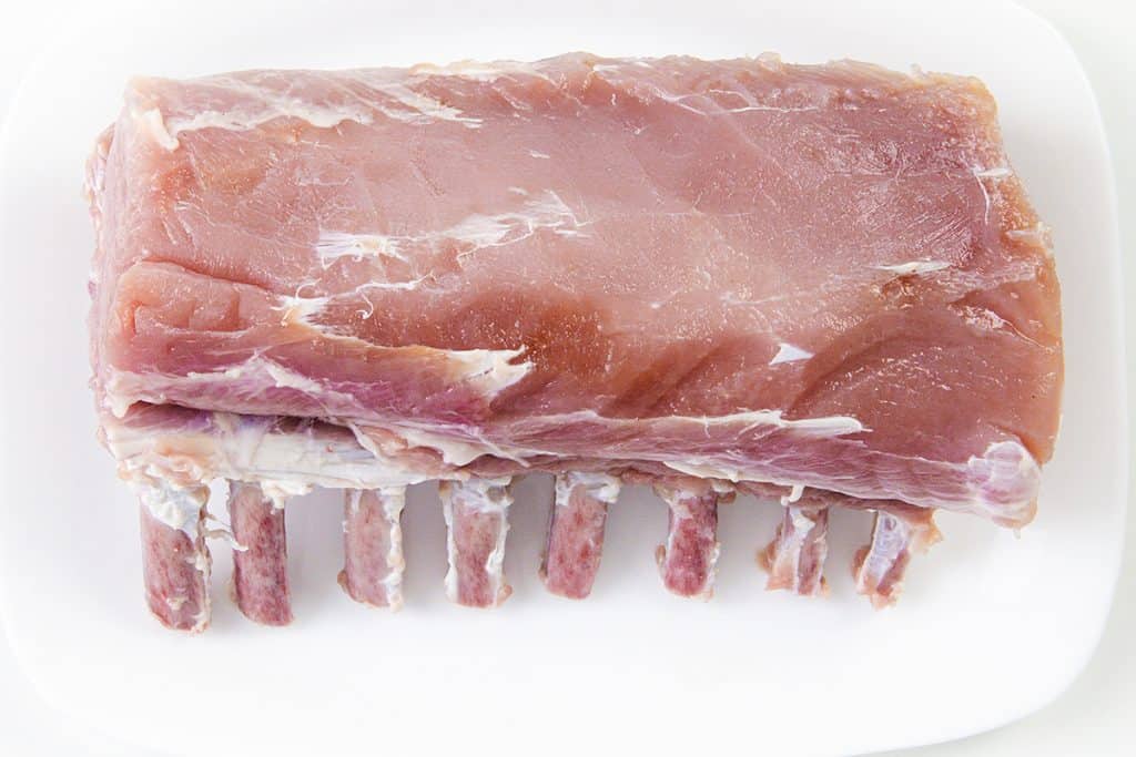 A raw Pork Rib Roast with the rib bones exposed at the bottom, placed on a white plate—perfect for preparing with a savory Garlic Herb Crust.