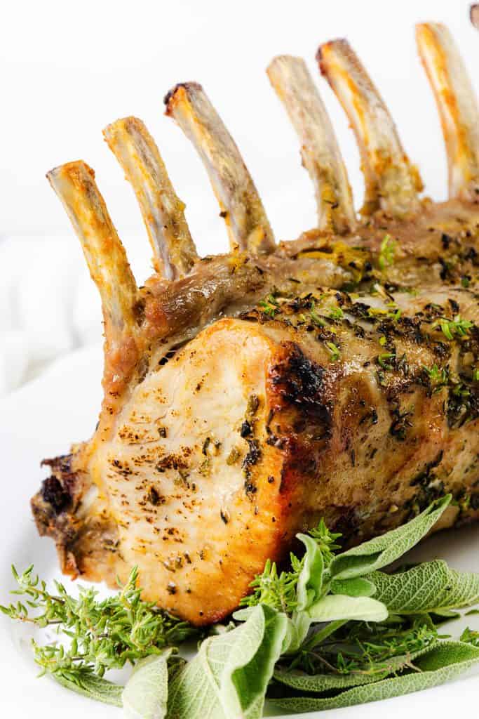 A close-up of a brined pork rib roast with a garlic herb crust on a white plate, garnished with fresh sprigs of sage and thyme.