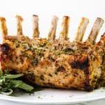 Herb-crusted Pork Rib Roast with exposed bones sits on a white platter, garnished with fresh green herbs. Plates and forks are in the background, resting on a white surface.