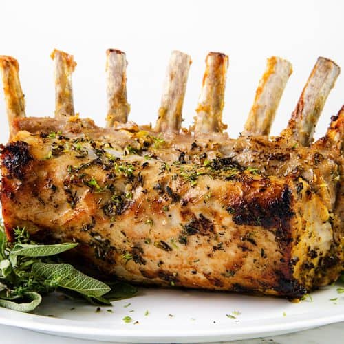 Herb-crusted Pork Rib Roast with exposed bones sits on a white platter, garnished with fresh green herbs. Plates and forks are in the background, resting on a white surface.