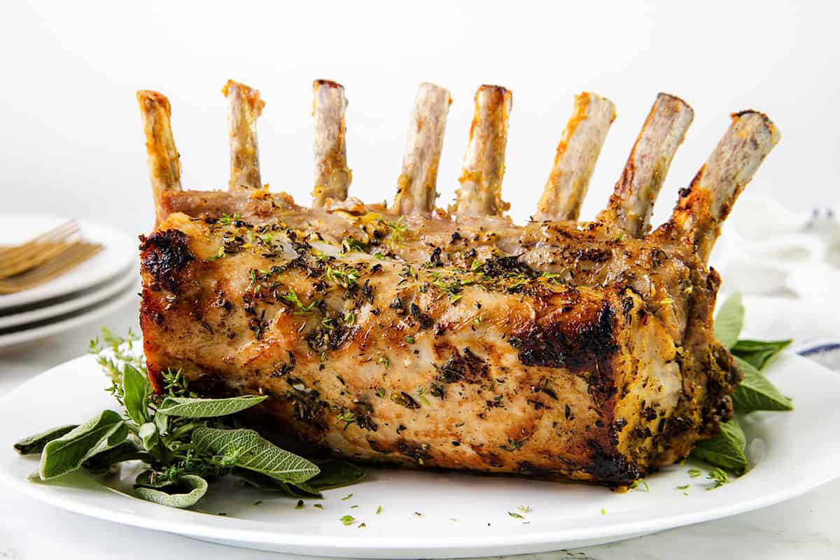 Herb-crusted Pork Rib Roast with exposed bones sits on a white platter, garnished with fresh green herbs. Plates and forks are in the background, resting on a white surface.