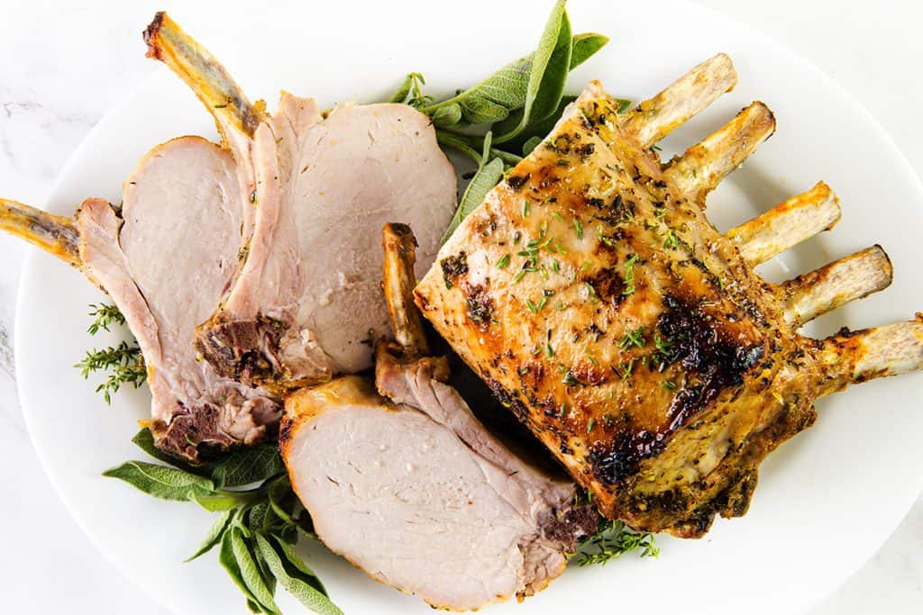 A cooked Pork Rib Roast with a golden garlic herb crust sits on a white plate, garnished with fresh herbs. Two slices have been cut and arranged beside the rack, revealing the juicy, tender meat inside.