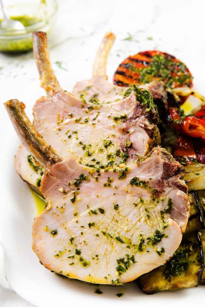Three cooked pork chops with a garlic herb crust are served on a white plate alongside grilled vegetables, including tomatoes and eggplant. The dish is drizzled with a vibrant green herb sauce.