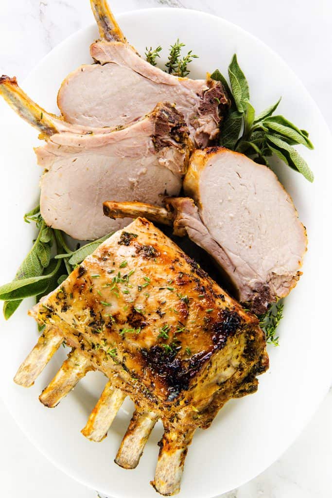 A white plate with a Garlic Herb Crust Pork Rib Roast, partially sliced into thick pieces and garnished with fresh herbs like sage and thyme. The meat appears juicy and cooked to a golden brown.