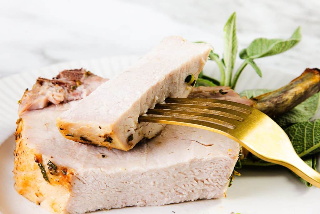 A close-up of a cooked pork chop with a savory garlic herb crust on a white plate, with two slices stacked and one held by a gold fork. Fresh sage leaves are placed beside the pork as garnish.