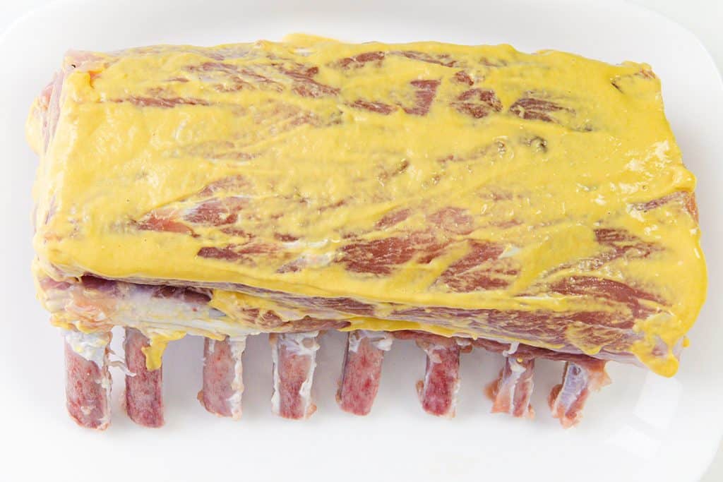 A raw pork rib roast coated with a thick layer of yellow mustard sits on a white surface, ready to become a flavorful garlic herb crust masterpiece.