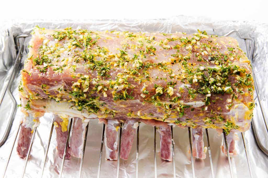 A raw rack of porkcoated with a flavorful garlic herb crust—made from chopped herbs, garlic, and spices—rests on a foil-lined roasting rack, ready to be cooked to perfection.