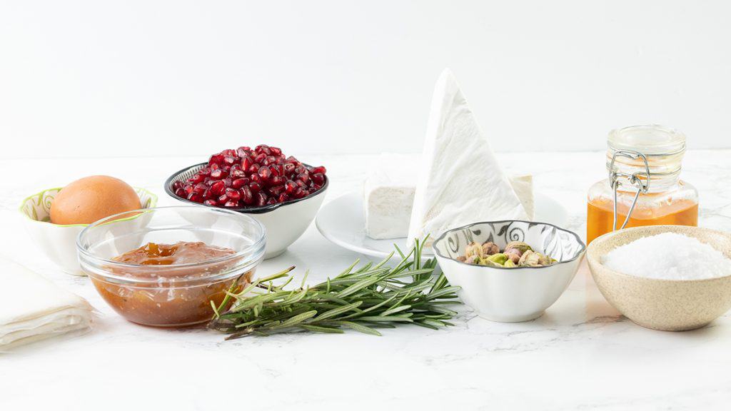 A variety of ingredients on a white surface, including a bowl of pomegranate seeds, wedge of Brie cheese, egg, rosemary sprigs, pistachios, coarse salt, and Fig Jam—perfect for assembling a Puff Pastry Christmas Wreath.
