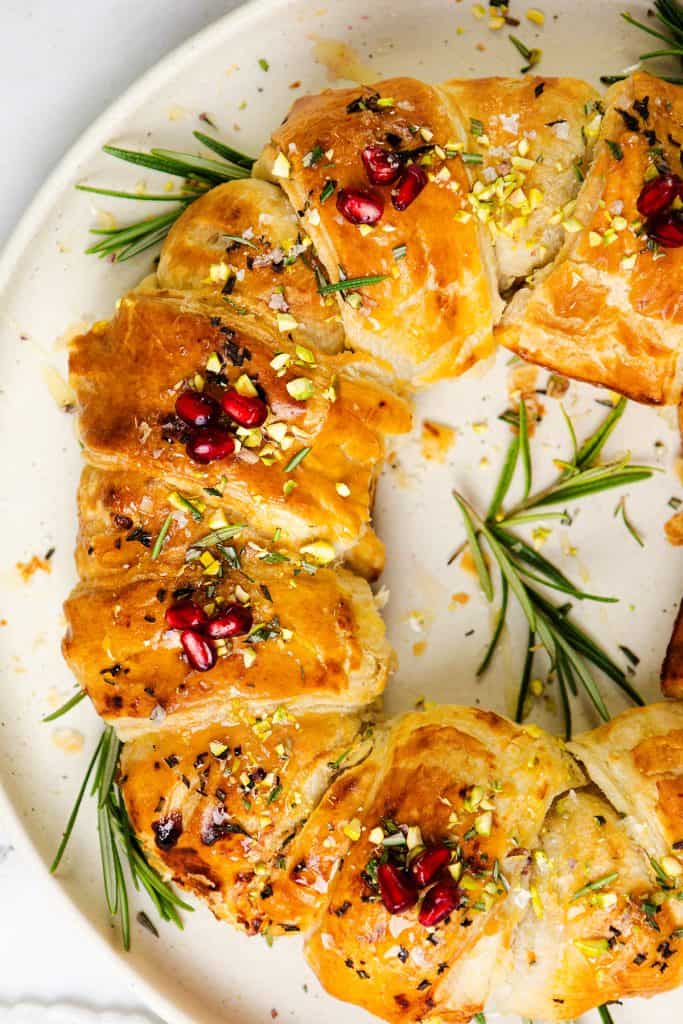 A golden, flaky wreath of brie in puff pastry with jam, topped with pomegranate seeds, chopped pistachios, sea salt, and fresh rosemary sprigs, arranged beautifully on a round white plate.