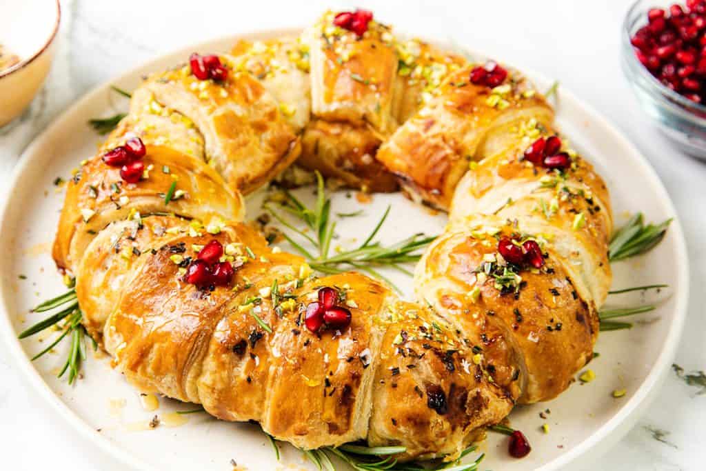 A golden-brown Puff Pastry Christmas Wreath is garnished with pomegranate seeds, chopped nuts, brie, and rosemary sprigs, arranged on a round white plate. A bowl of pomegranate seeds sits nearby.
