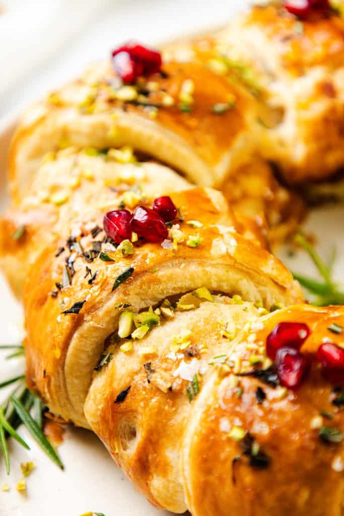 A close-up of a golden-brown, braided bread topped with chopped pistachios, fresh rosemary, coarse salt, red pomegranate seeds, and a hint of fig jam. The bread looks glossy and freshly baked.