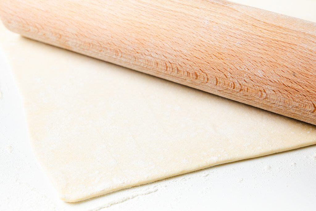 A wooden rolling pin rolling out a sheet of dough on a lightly floured white surface, perfect for shaping a Puff Pastry Christmas Wreath.