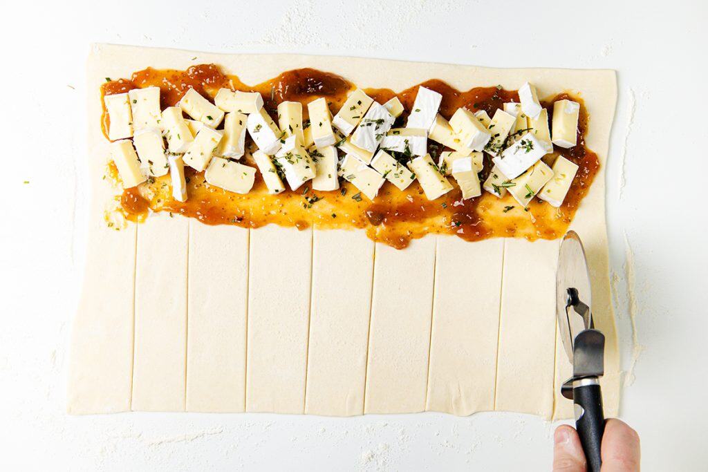 A hand uses a pastry cutter to slice rectangular strips into puff pastry topped with brie, herbs, and a layer of fig jam, preparing a savory treat on a white surface.
